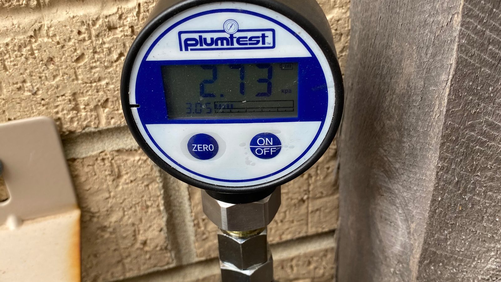 A Plumtest digital pressure gauge fitted to a pipe against a brick wall