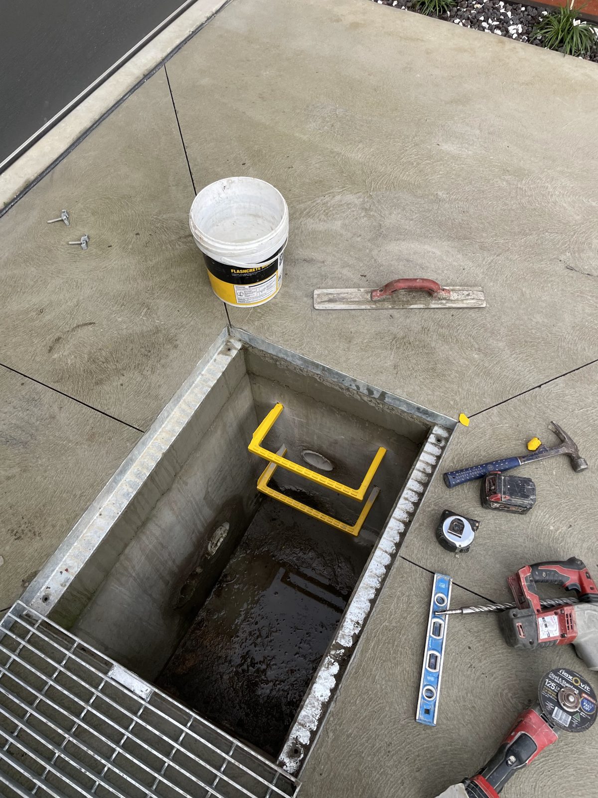 Looking down into a galvanised pit set into concrete, with a steel grate, level, hammer and drill laid out around it