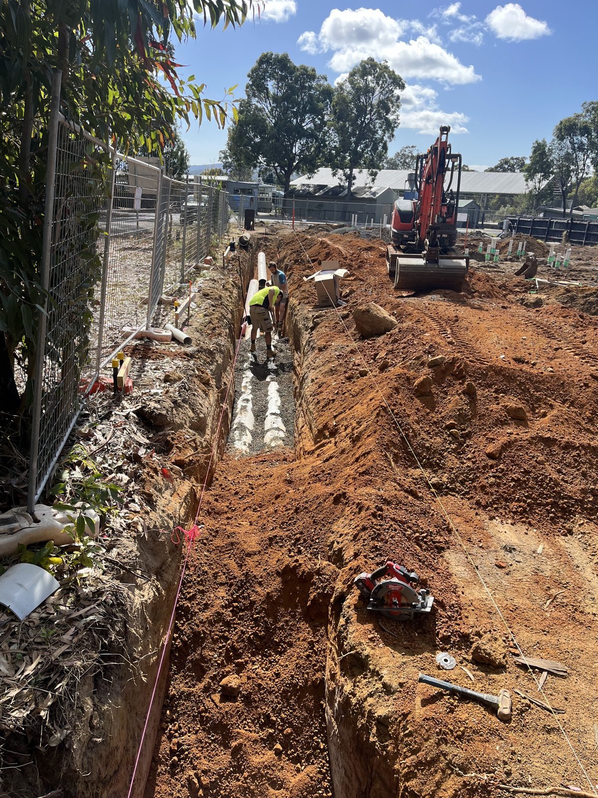 A long trench in red clay on a building site, pipe laid on a gravel bed, workers in hi-vis and an excavator beyond