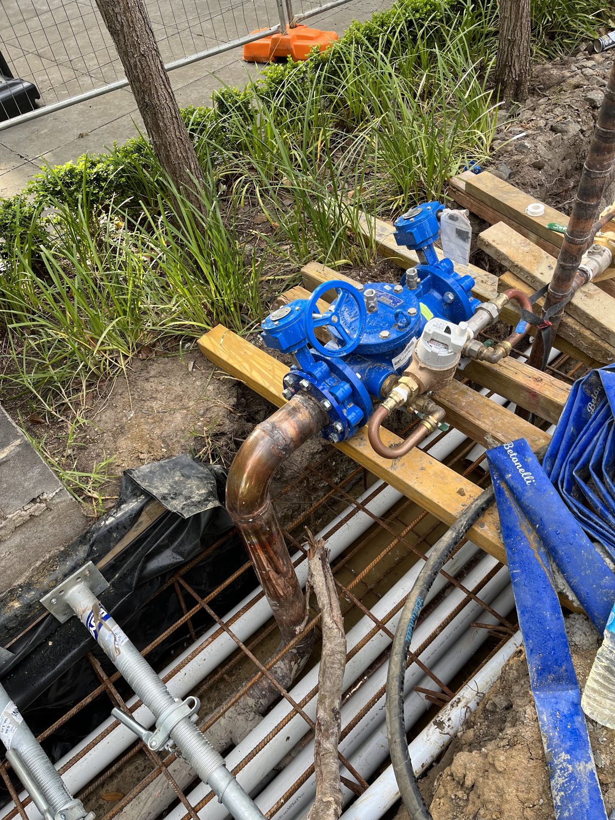 Blue gate valves and a water meter on copper pipework, set on timber beside a garden bed on a building site
